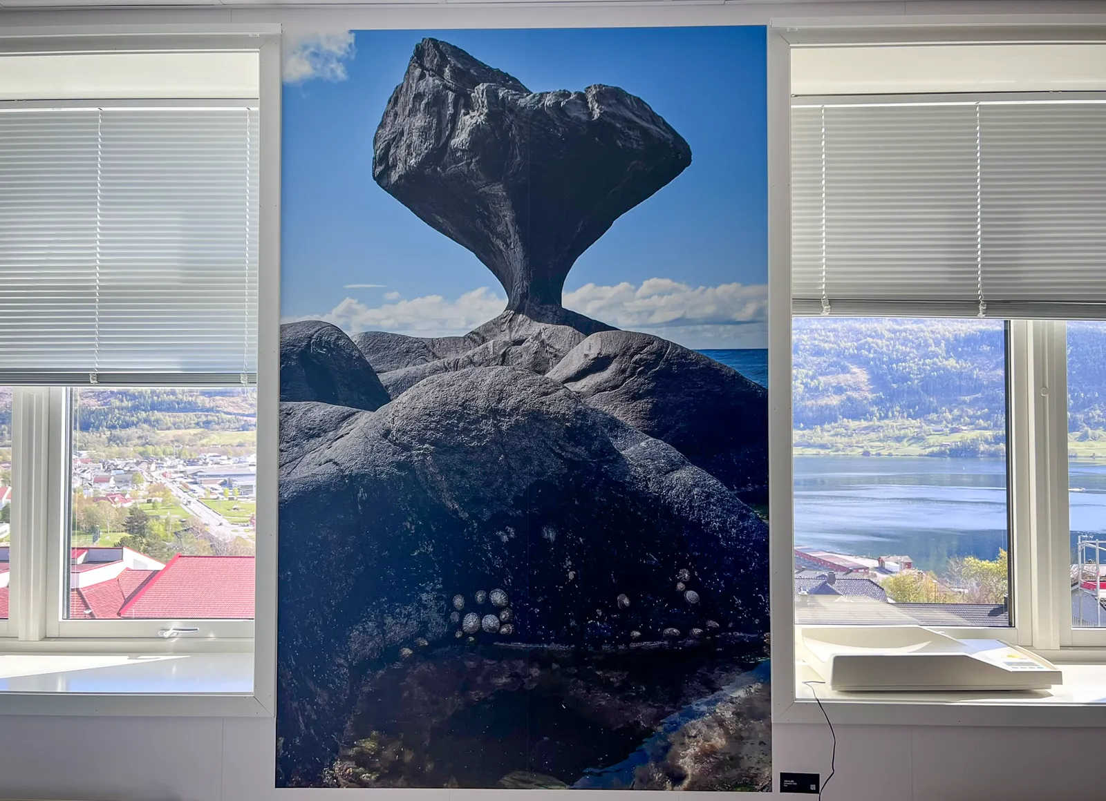 Fototapet mellom to vinduer på eit barselrom ved Nordfjord sjukehus. Tapeten viser ein skulptur av ein stein mot blå himmel.