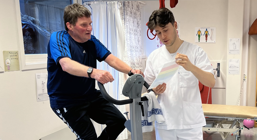 Ein mann sit på ein ergometersykkel i treningskle mens ein mann i kvit uniform viser han eit skjema
