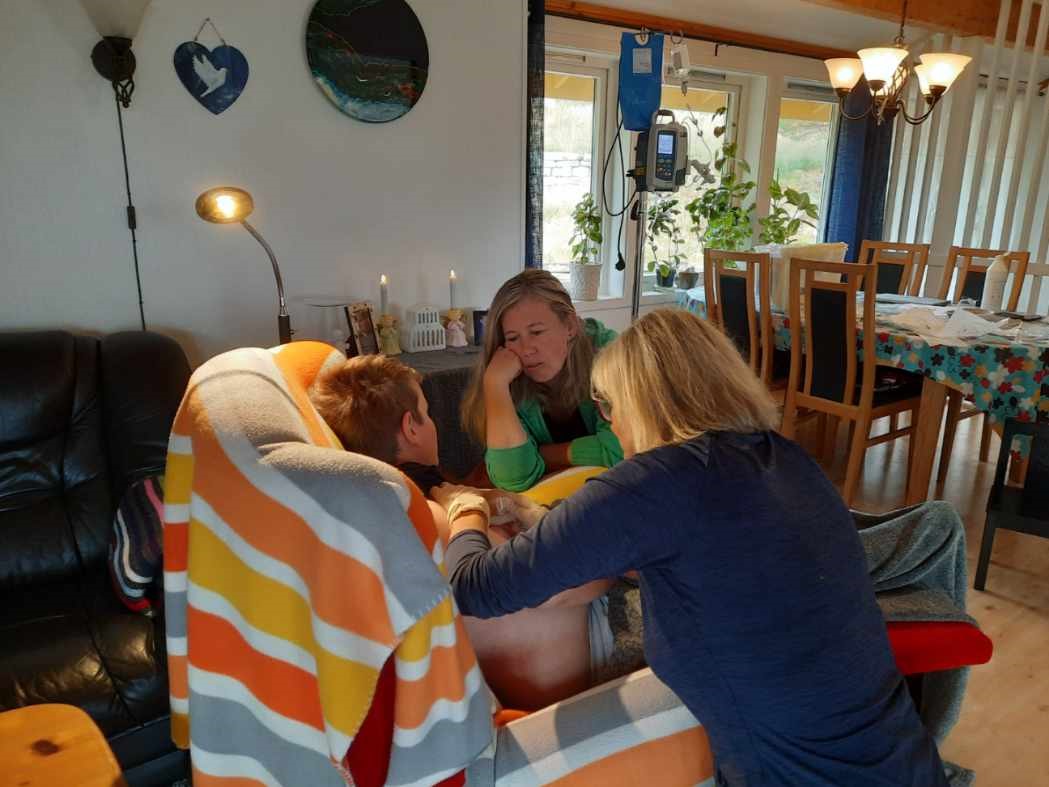 et barn sitter i en stol og får behandling av en sykepleier i hjemmet. En kvinne med grønn genser sitter ved siden av barnet.