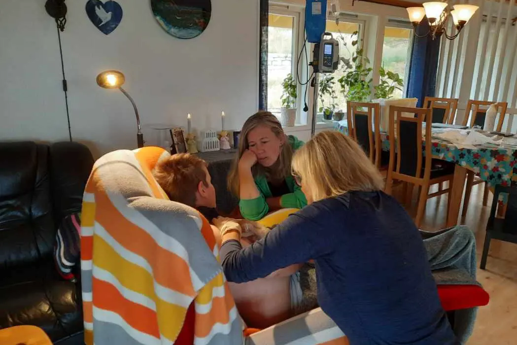 et barn sitter i en stol og får behandling av en sykepleier i hjemmet. En kvinne med grønn genser sitter ved siden av barnet.