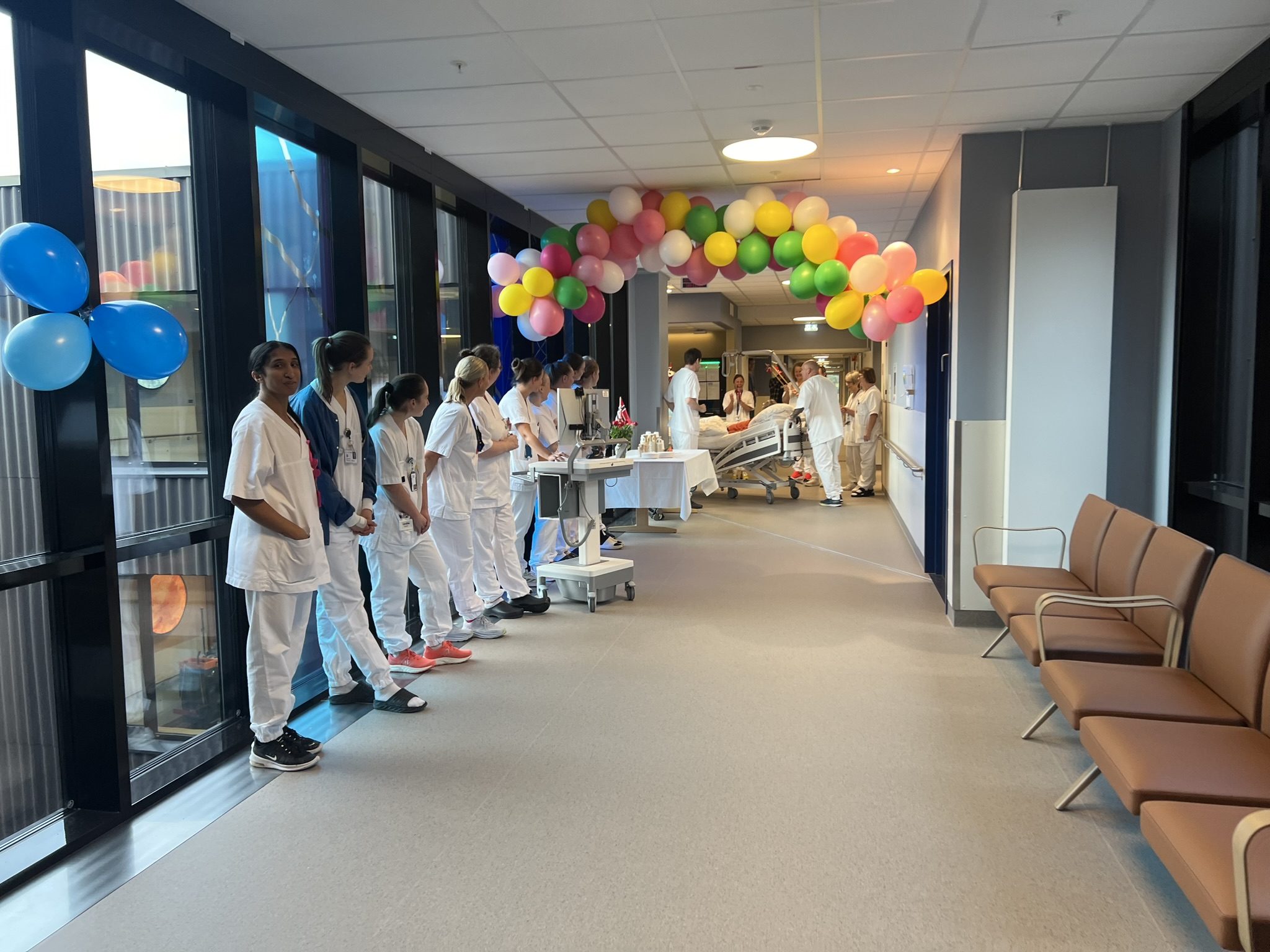 En gruppe ansatte i hvite uniformer som står i en gang med ballonger, mens den første pasienten trilles inn i Livabygget.