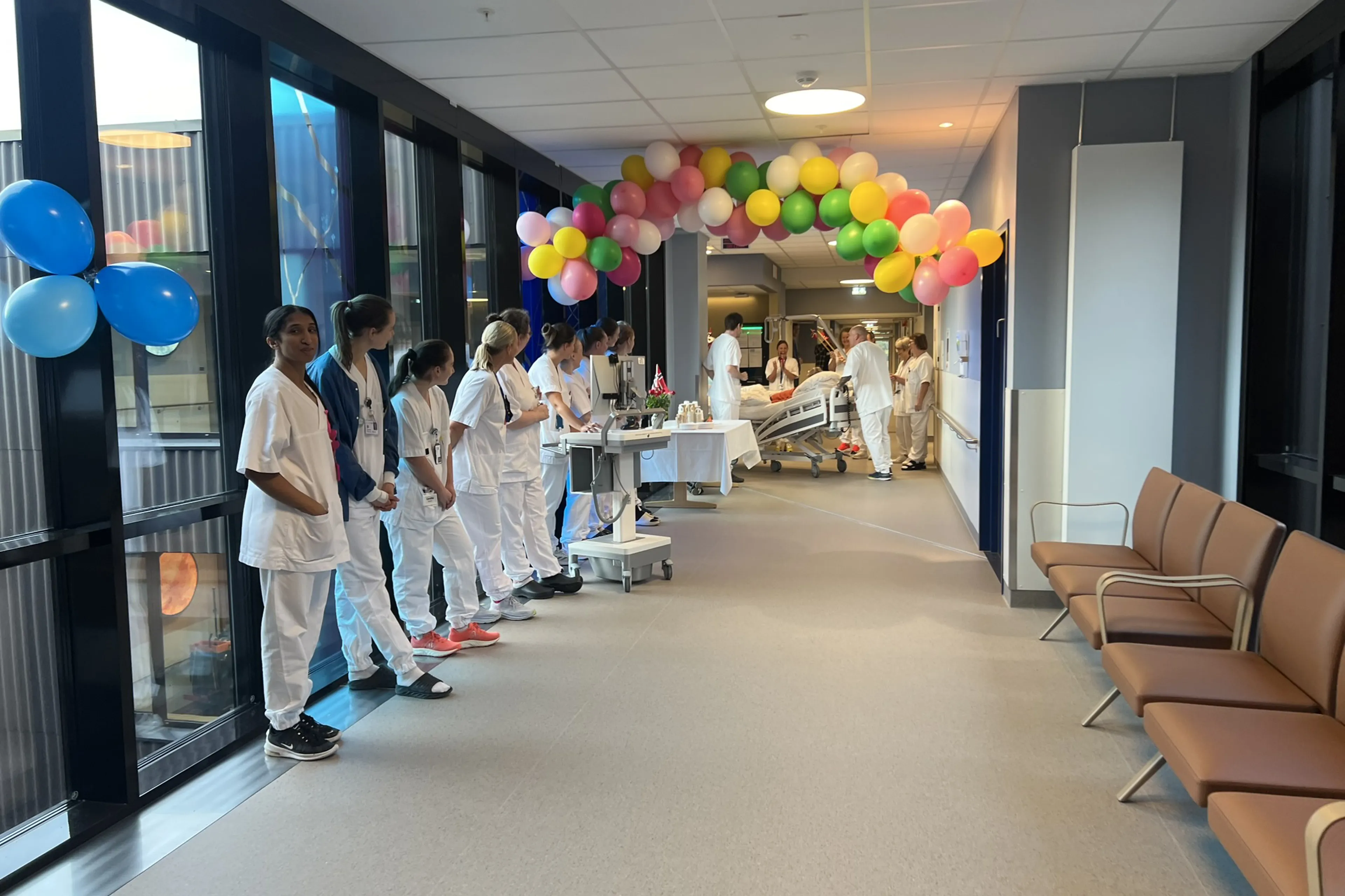 En gruppe ansatte i hvite uniformer som står i en gang med ballonger, mens den første pasienten trilles inn i Livabygget.