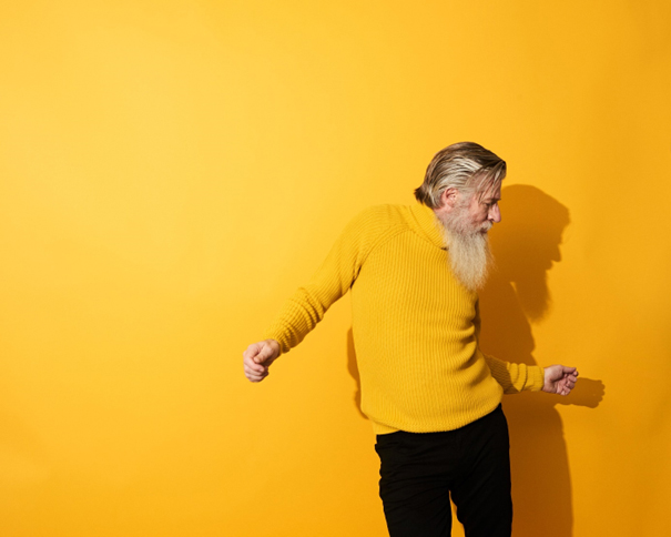 En eldre mann med skjegg som danser foran en gul vegg