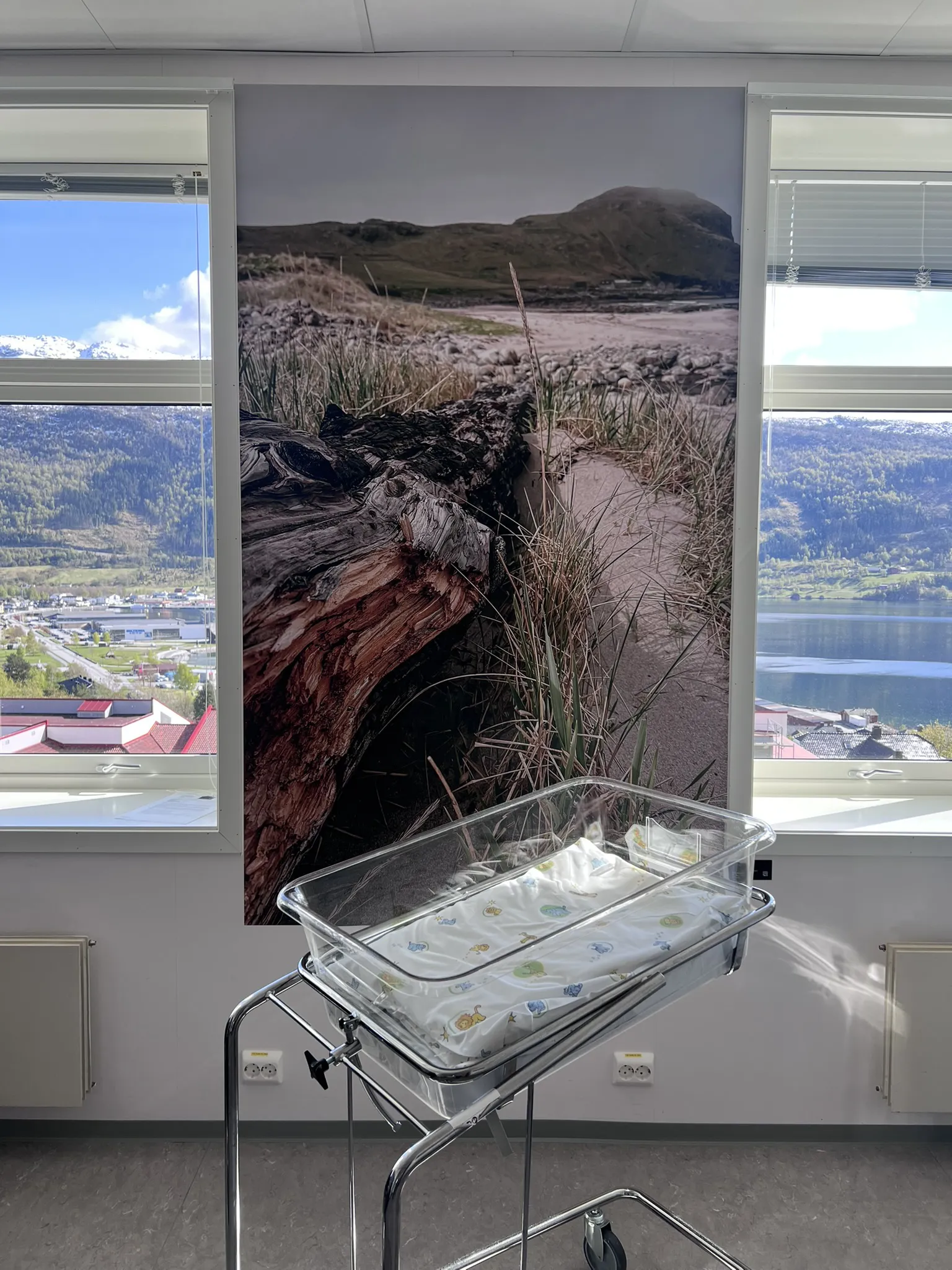 Fotokunst som viser ei strand inne på eit barselrom med ei barneseng framfor