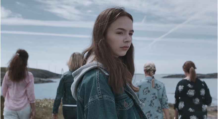 Ei ungjente med dongerijakke som står ute, med fleire ungdommar i bakgrunnen