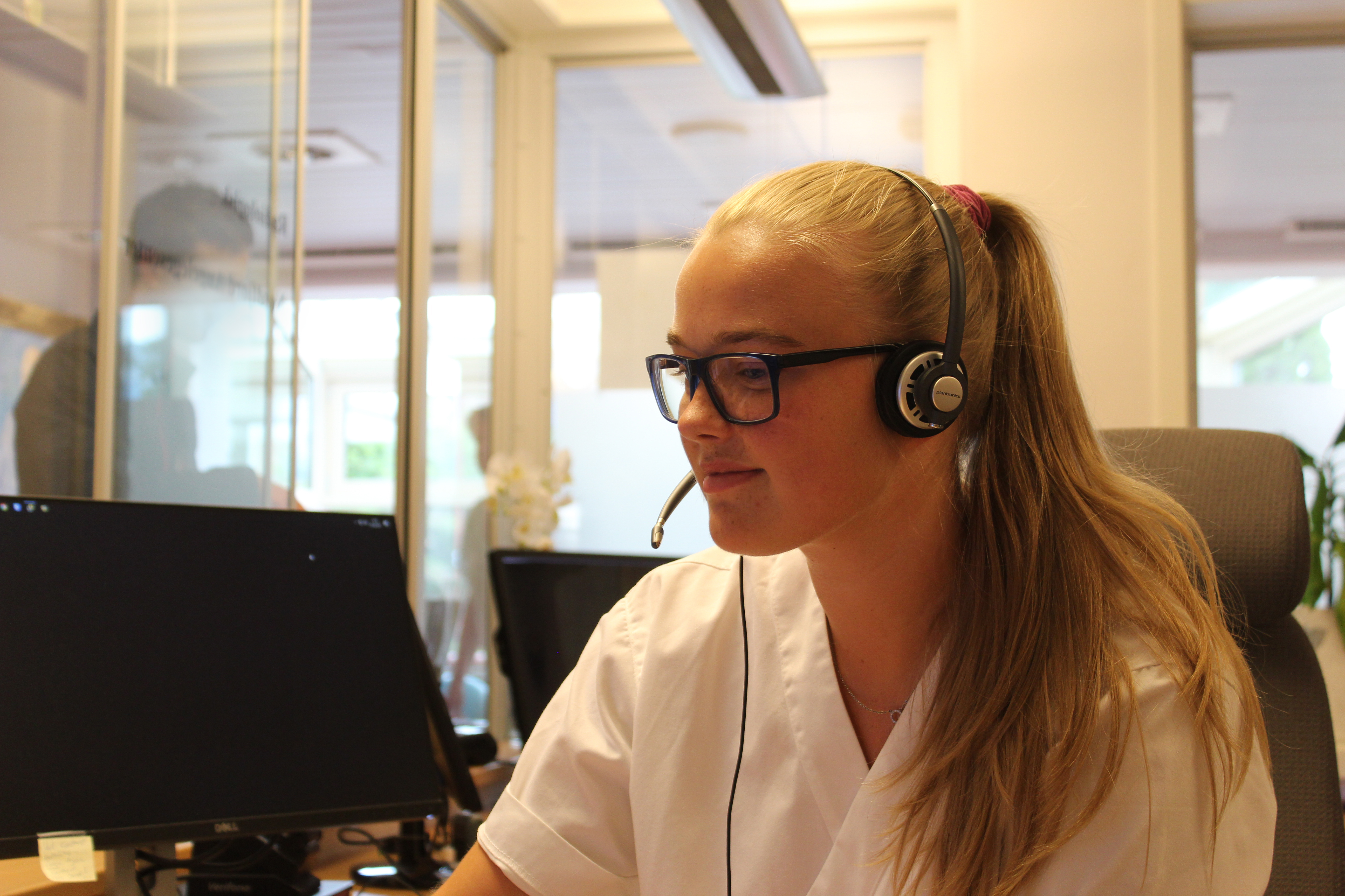 En ung kvinne i hvit uniform sitter foran en dataskjerm med hodetelefoner og mikrofon