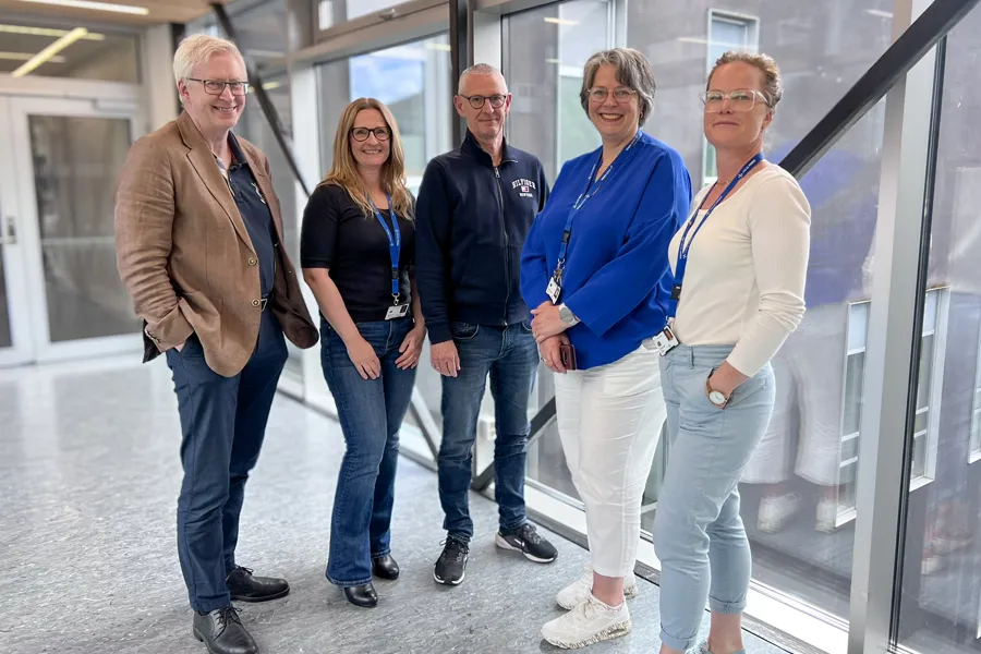 Fem personar som står i ein glasskorridor på sjukehuset.