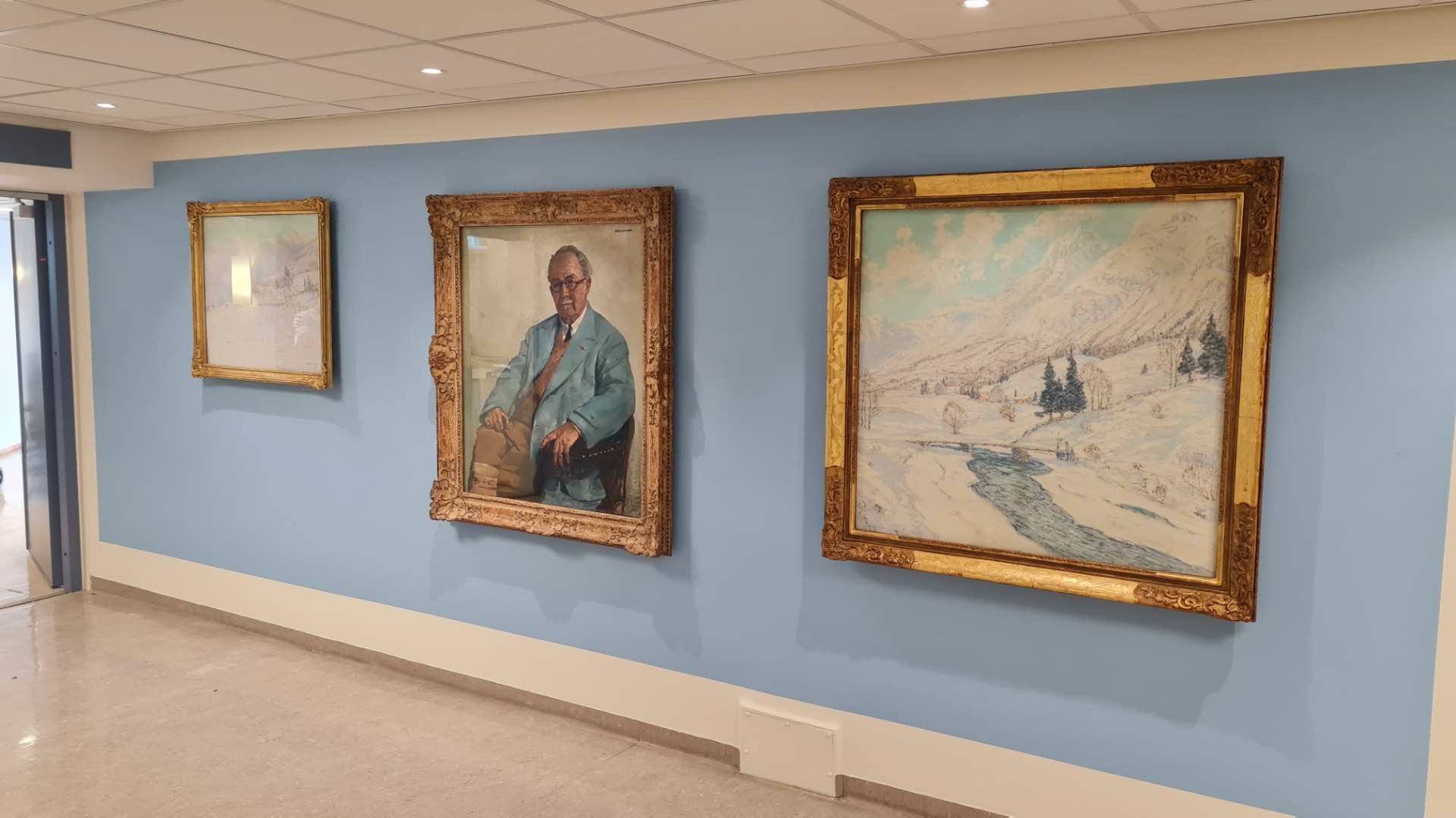 Tre måleri i gullrammer som heng på ein lyseblå vegg på sjukehuset
