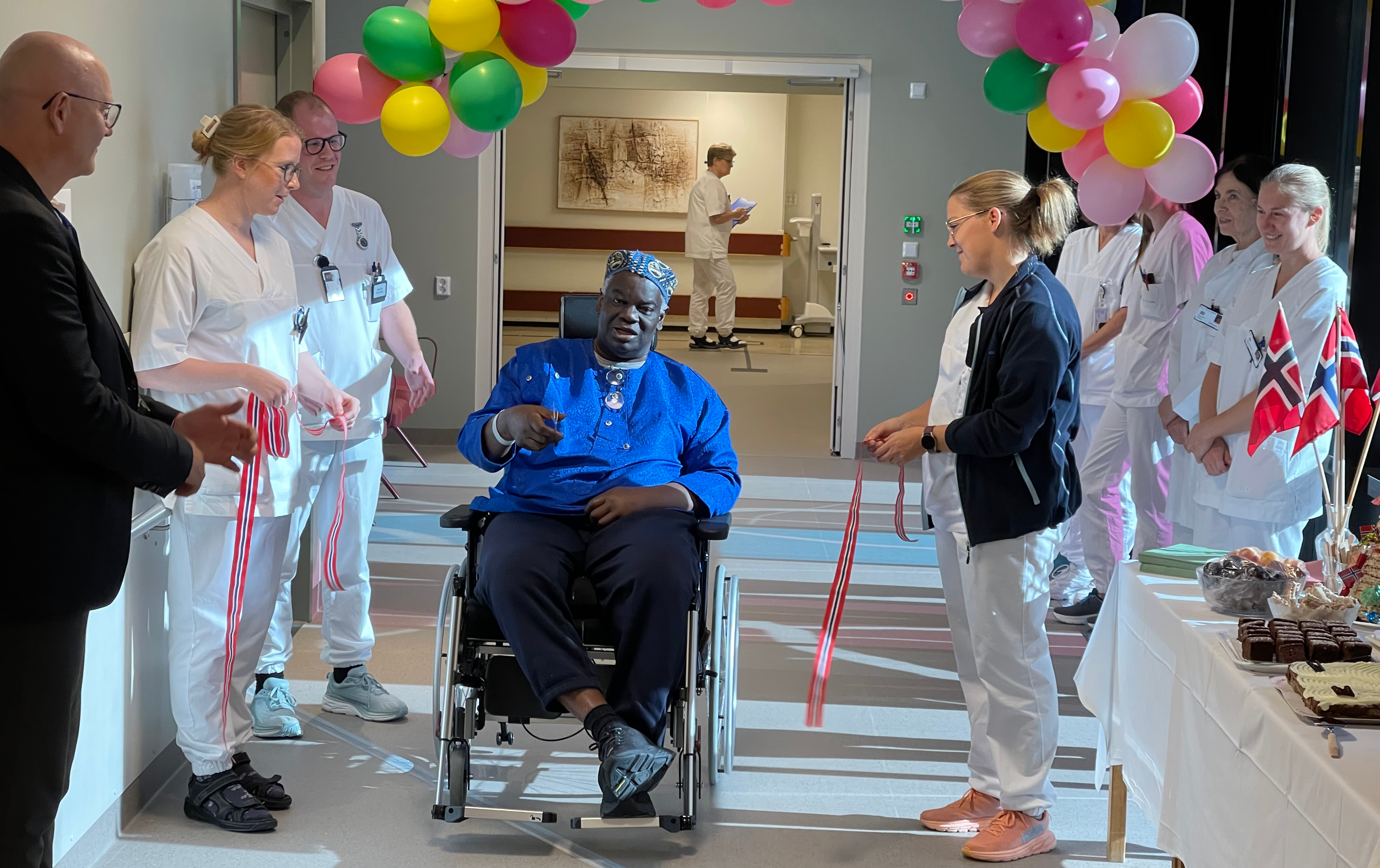 En mann i rullestol og en rekke helsepersonell i en gang med ballonger og flagg