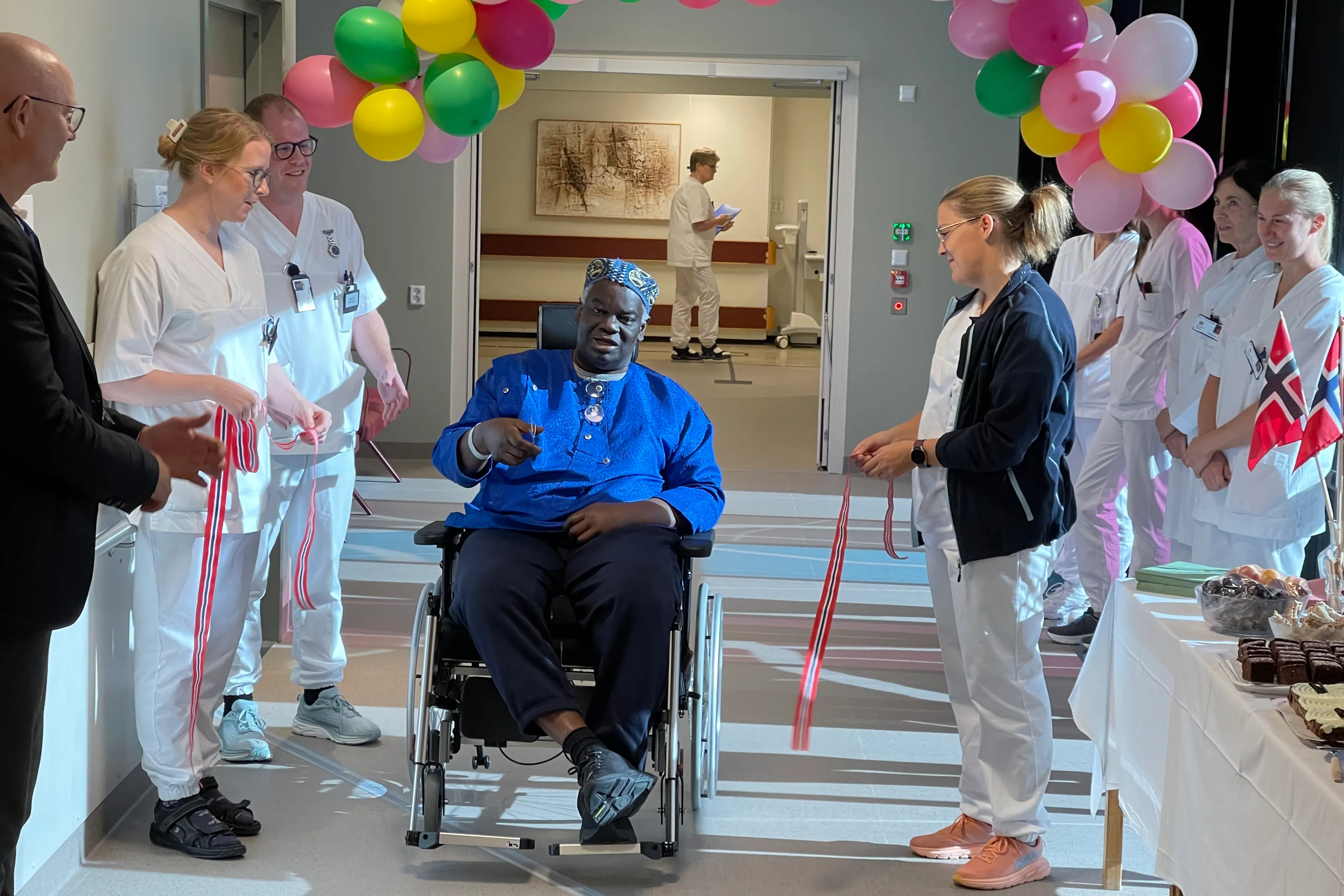 En mann i rullestol og en rekke helsepersonell i en gang med ballonger og flagg