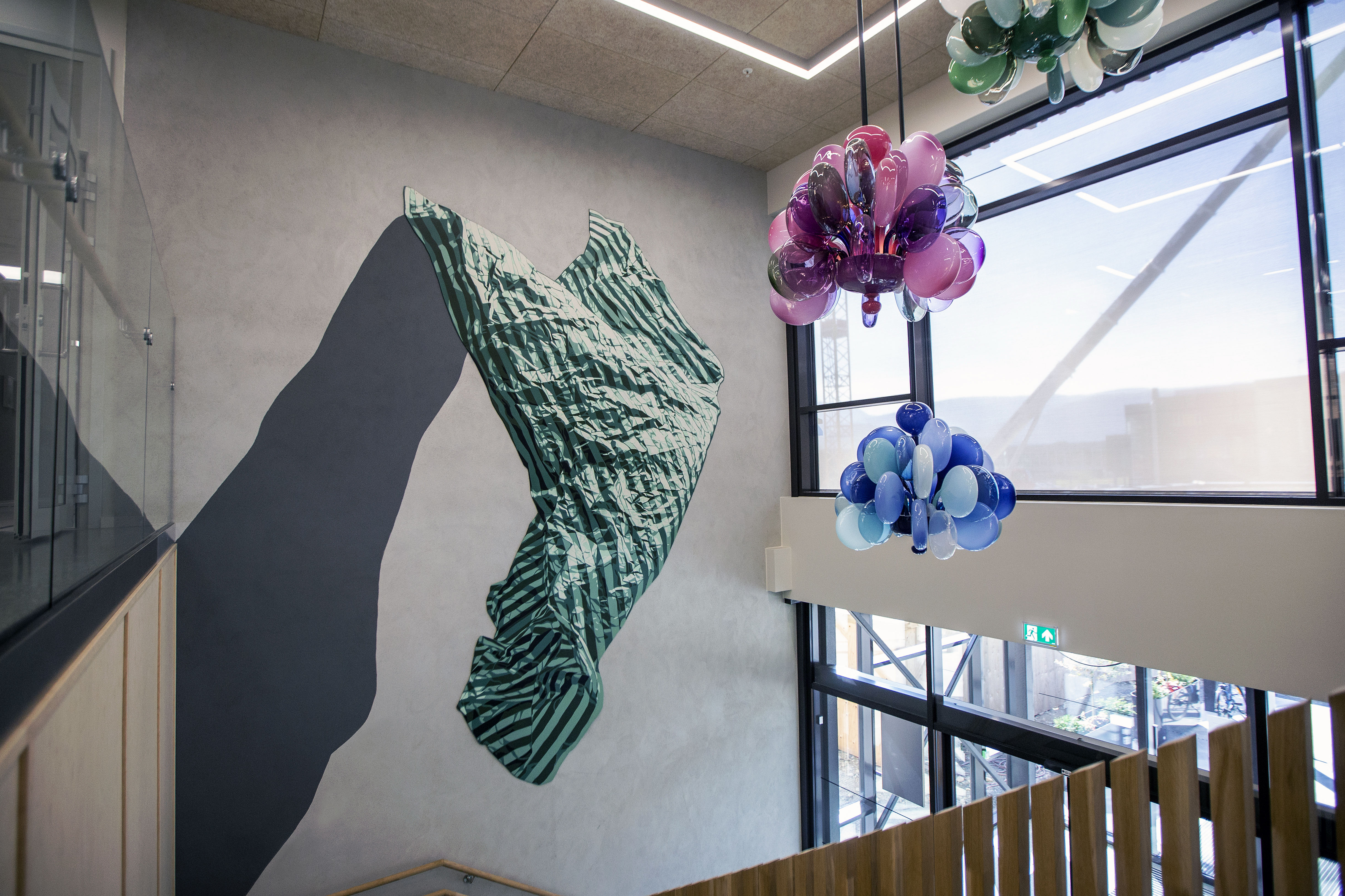 tre lysekruner i glas i rosa, blått og grønt, samt ein veggkonstruksjon som heng på ein betongvegg