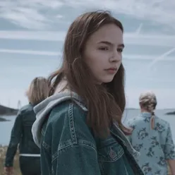 Ei ung jente med dongerijakke som står ute, med fleire ungdomar i bakgrunnen