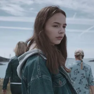 Ei ung jente med dongerijakke som står ute, med fleire ungdomar i bakgrunnen