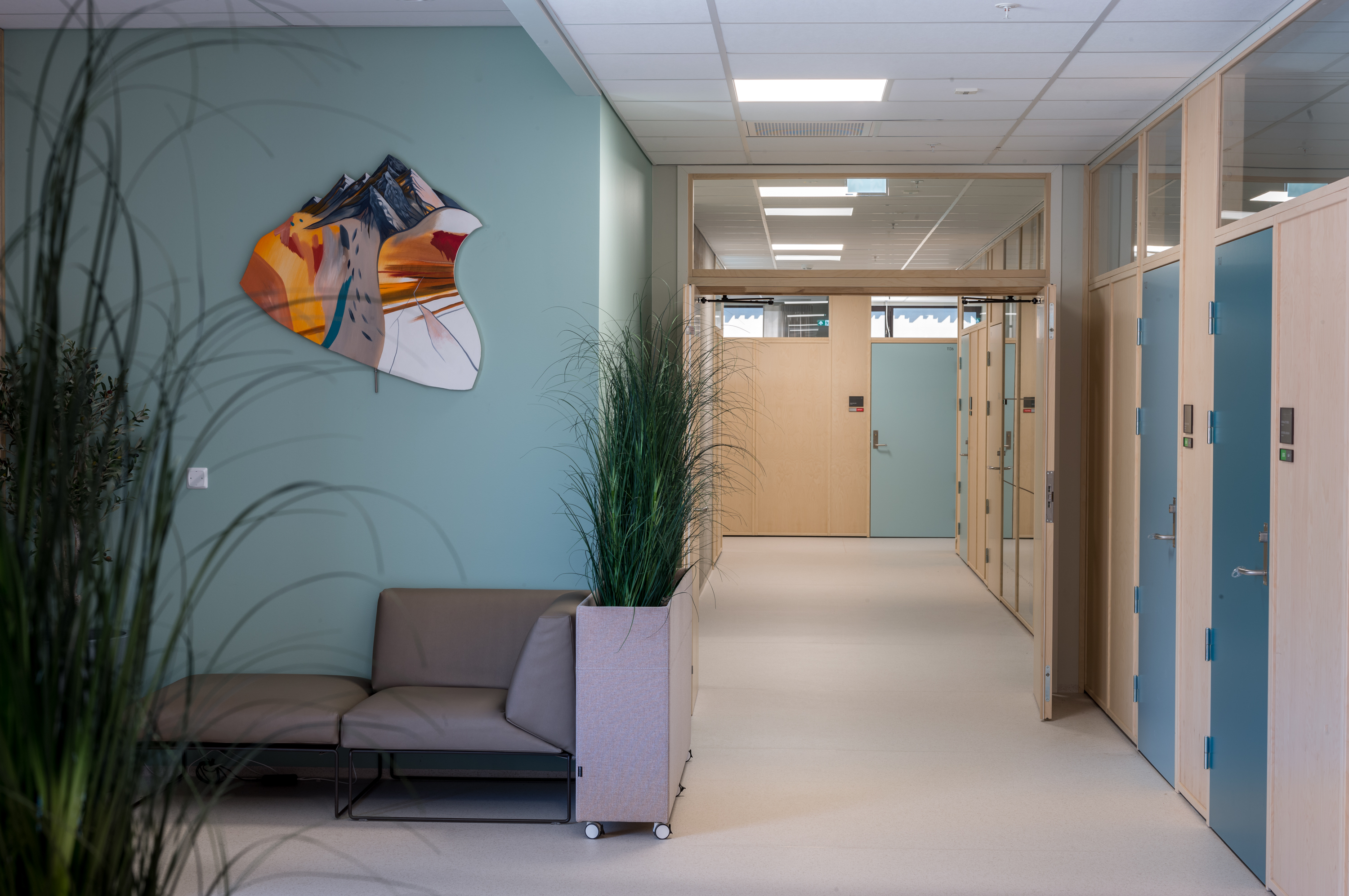 korridor med 2 grøne plantar, ein sjeselong med ein grøn vegg bak der det heng eit fargerikt kunstverk