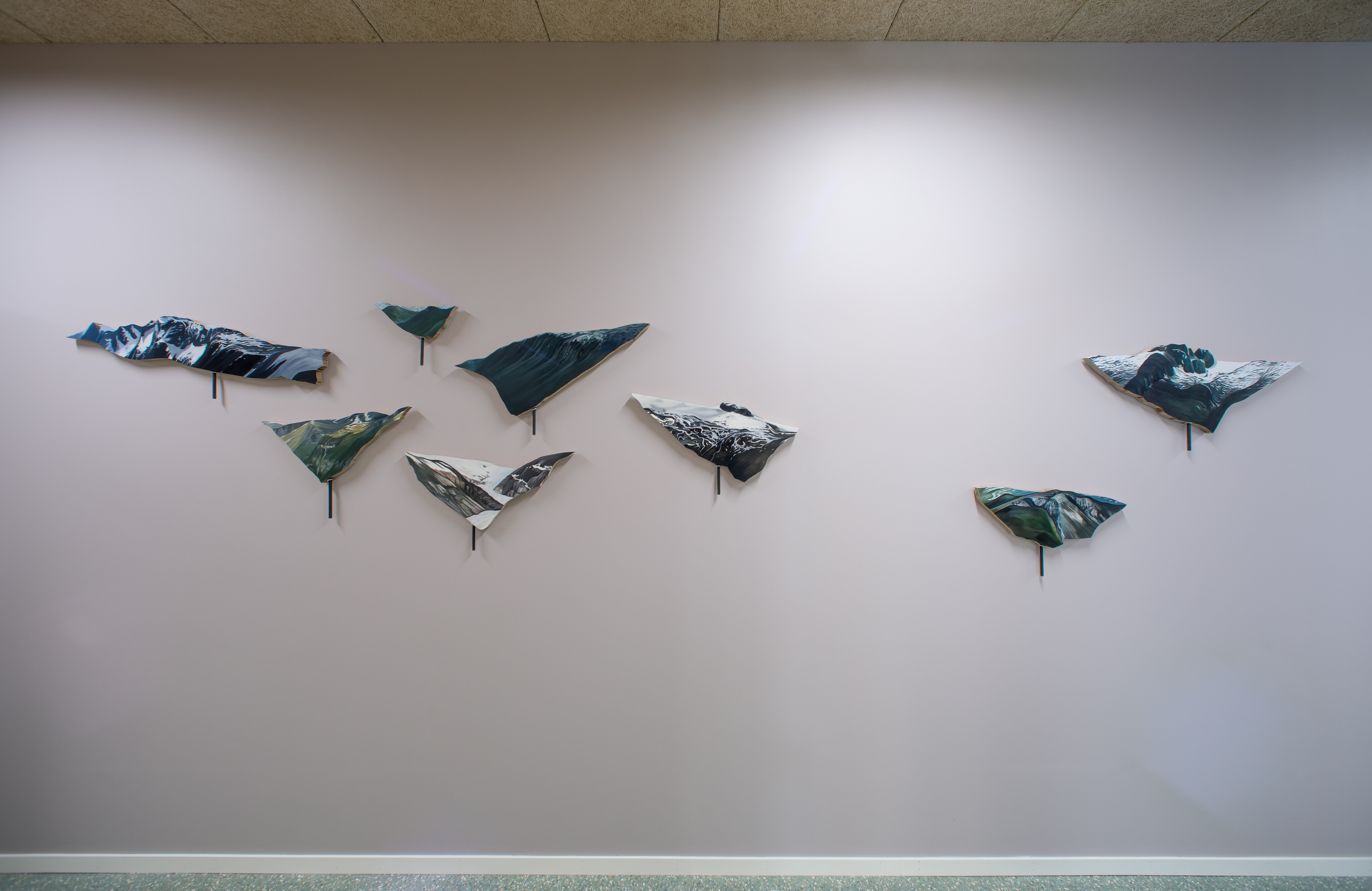 Ein kvit vegg med 8 kunstverk i ulike storleikar som heng på veggen og ser ut som fuglar. 