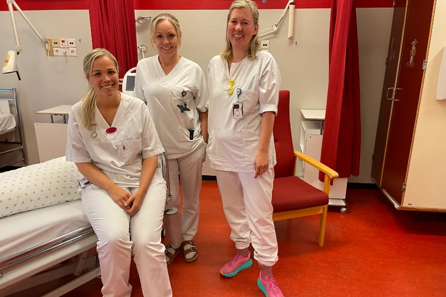 tre kvinnelege sjukepleiarar som er kledd i kvit uniform inne på eit pasientrom med raude gardiner og møblement.