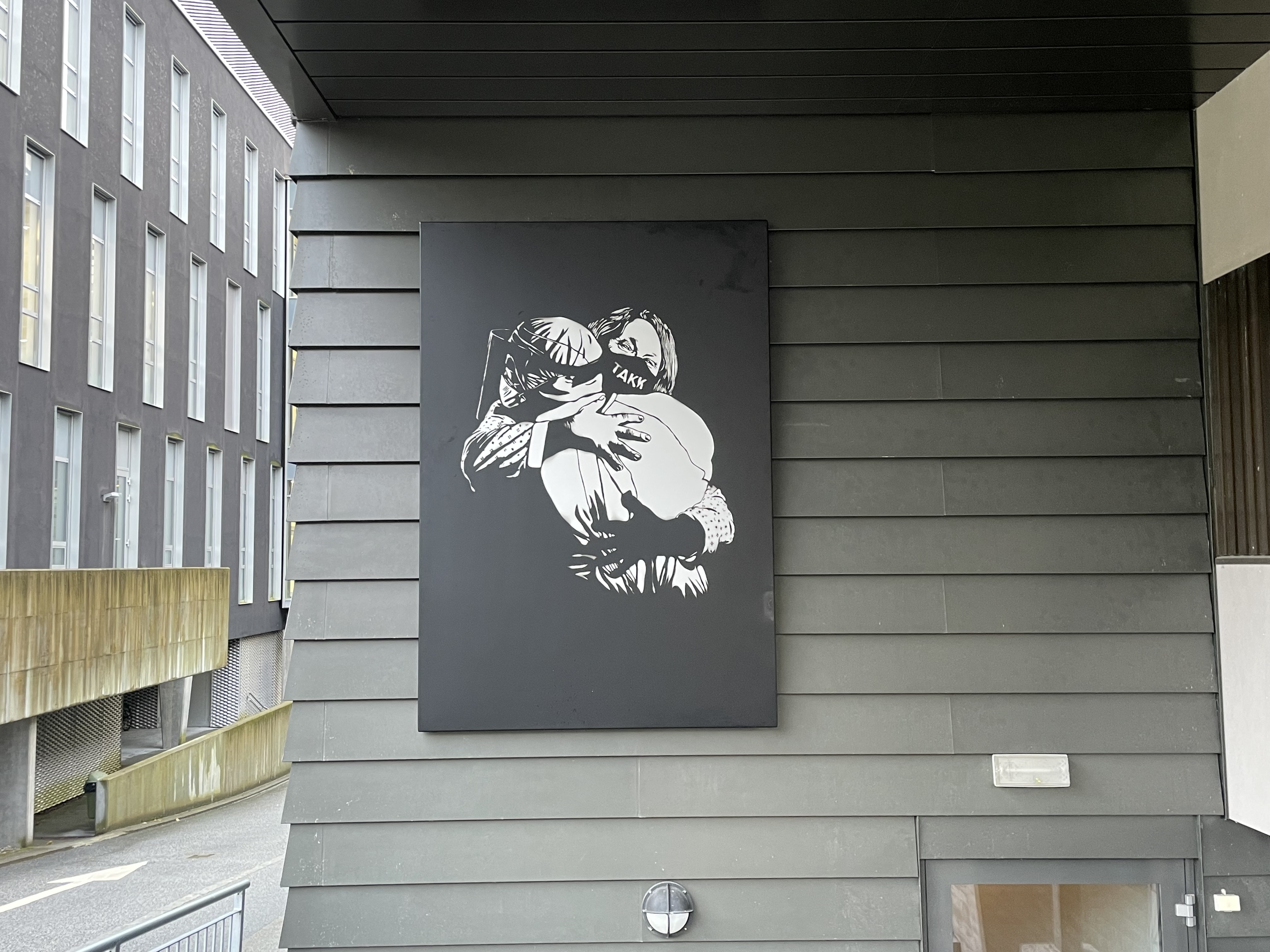 utandørs kunstverk som heng på ein gråmalt vegg av to personar som omfavnar kvarandre