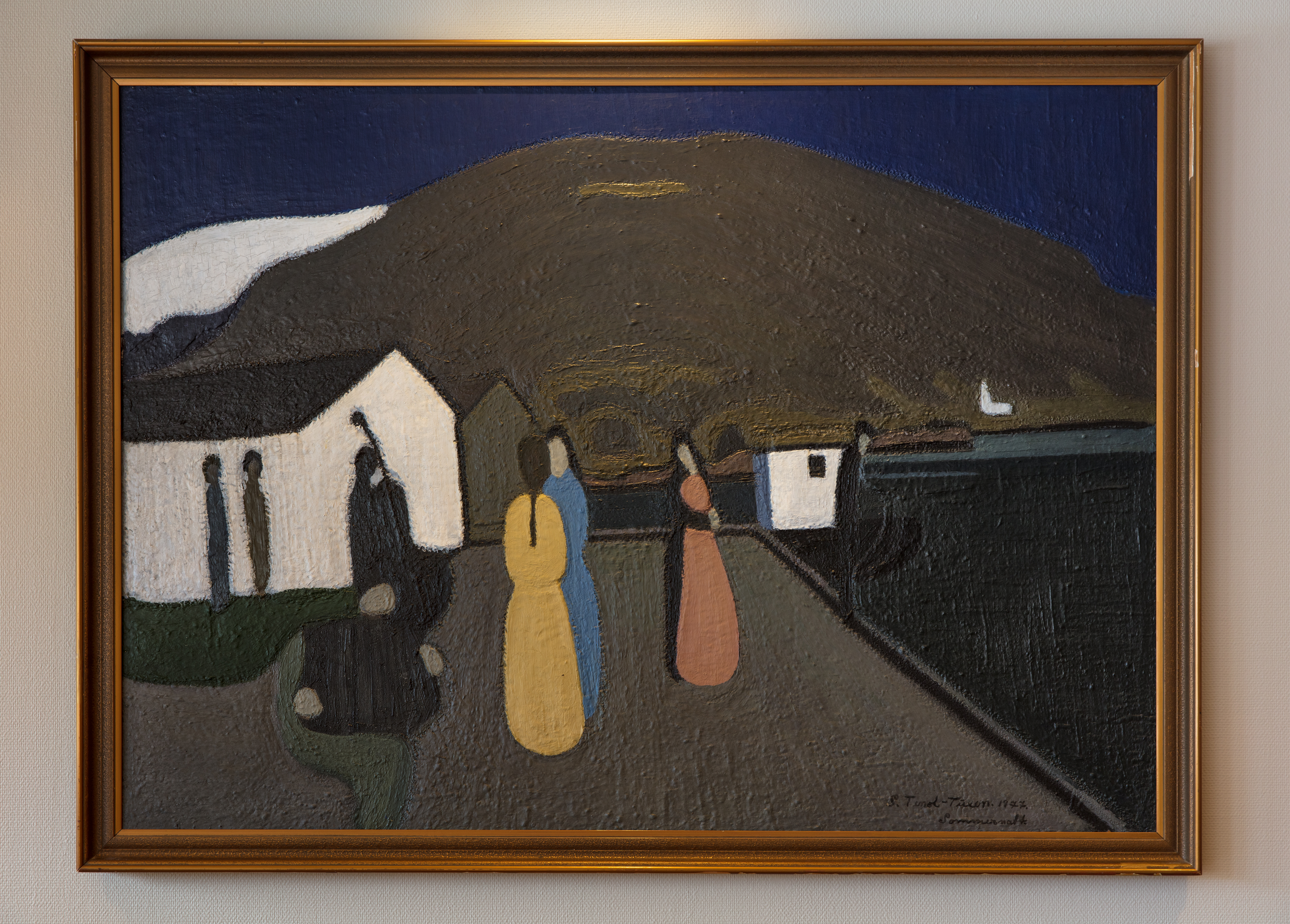 måleri i brun ramme som heng på ein vegg. Motivet er av tre kvinner i fargerike kjolar som står på ei brygge med fjord og fjell 