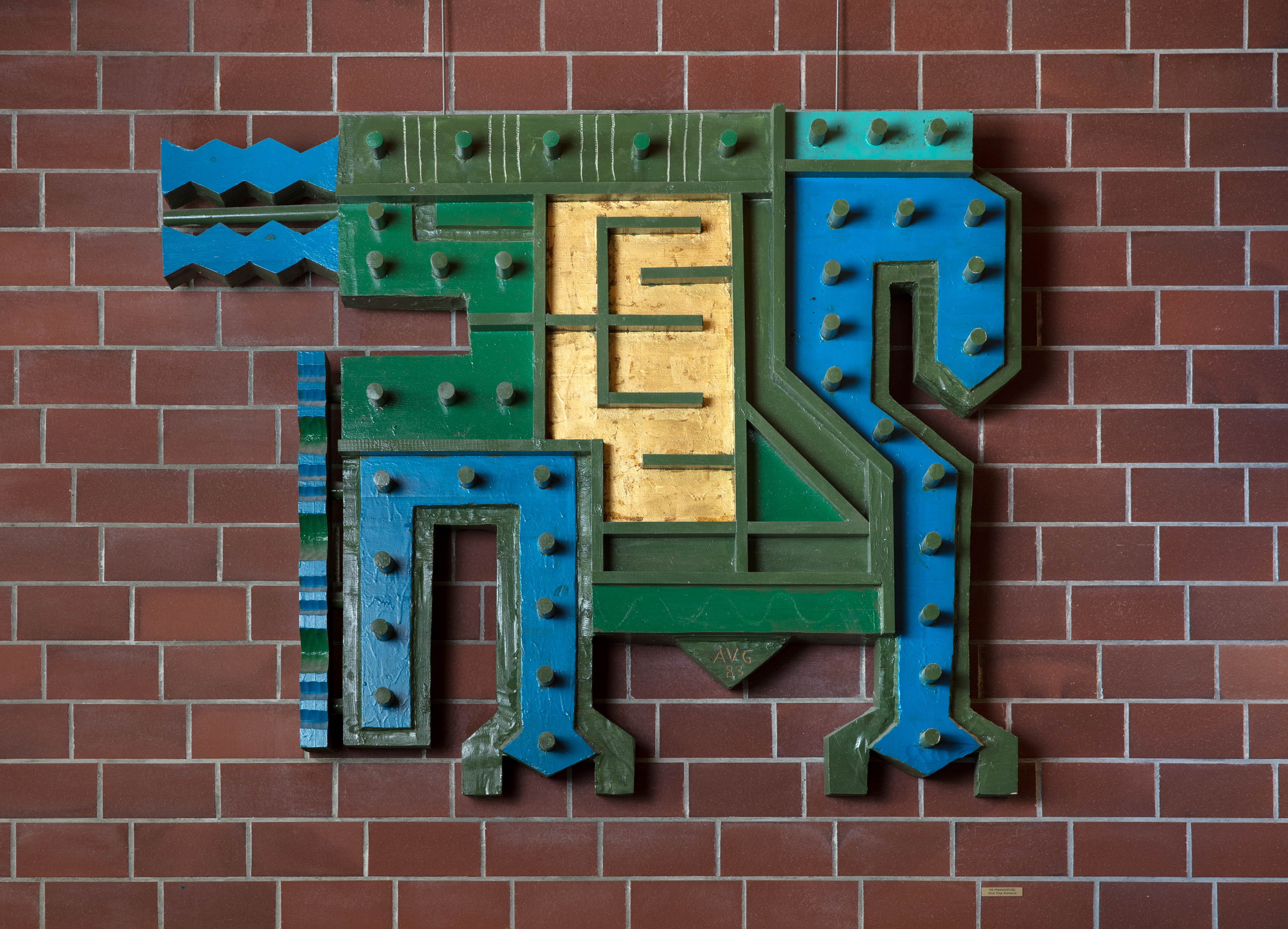 Raud mursteinsvegg med eit kunstverk i tre i fargane blå, grøn og gul som forstiller ein fugl